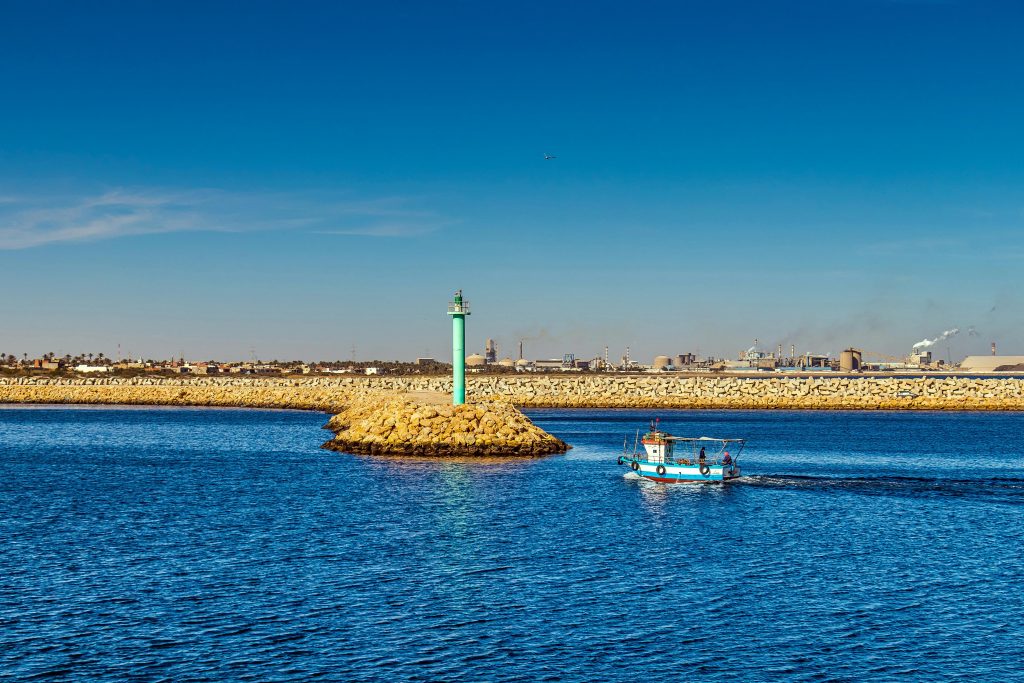 A small fishing boat sailing past a tall lighthouse in Gabès harbor, Tunisie.